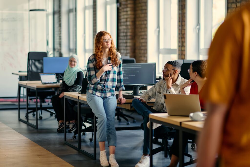 A group of five professionals engage in a conversation around desks with laptops and monitors, highlighting teamwork in a contemporary software development setting.